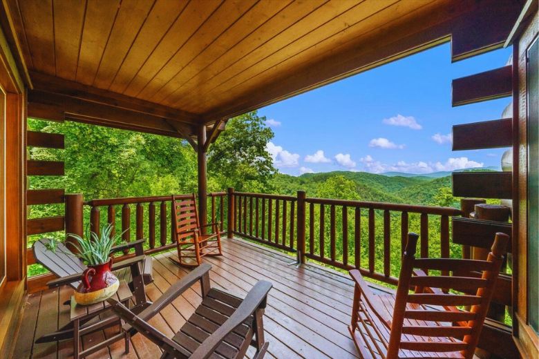 Covered wooden cabin deck with rocking chairs and a small table overlooking a lush green forest and distant mountain views under a bright blue sky with scattered clouds.
